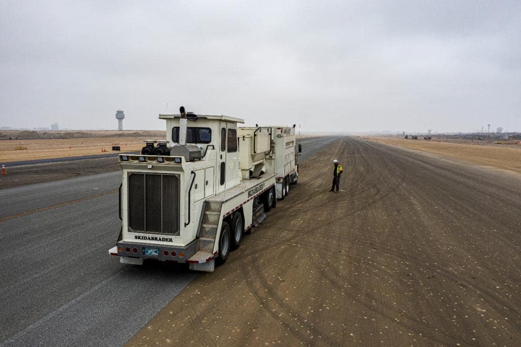 A large vehicle labeled "Skidabrader" parked on a wide, empty runway under a cloudy sky. A person in a safety vest stands nearby, emphasizing the vehicle's scale.