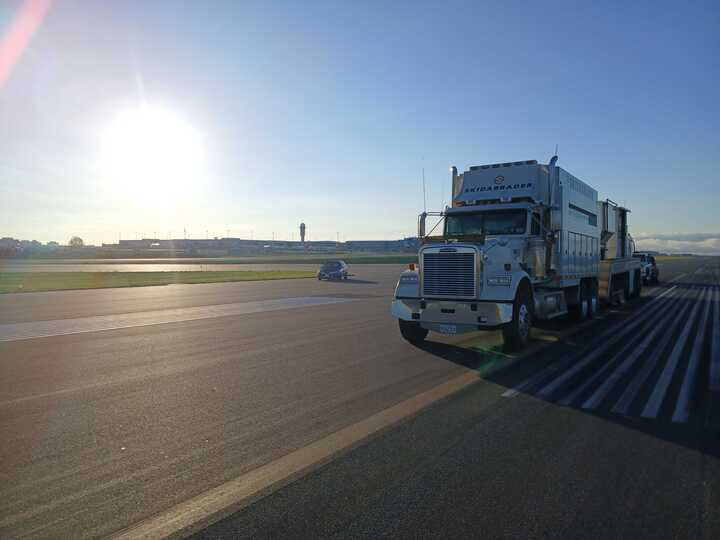 A large white industrial truck on a runway under a bright morning sun, casting long shadows. A control tower is visible in the distant background.