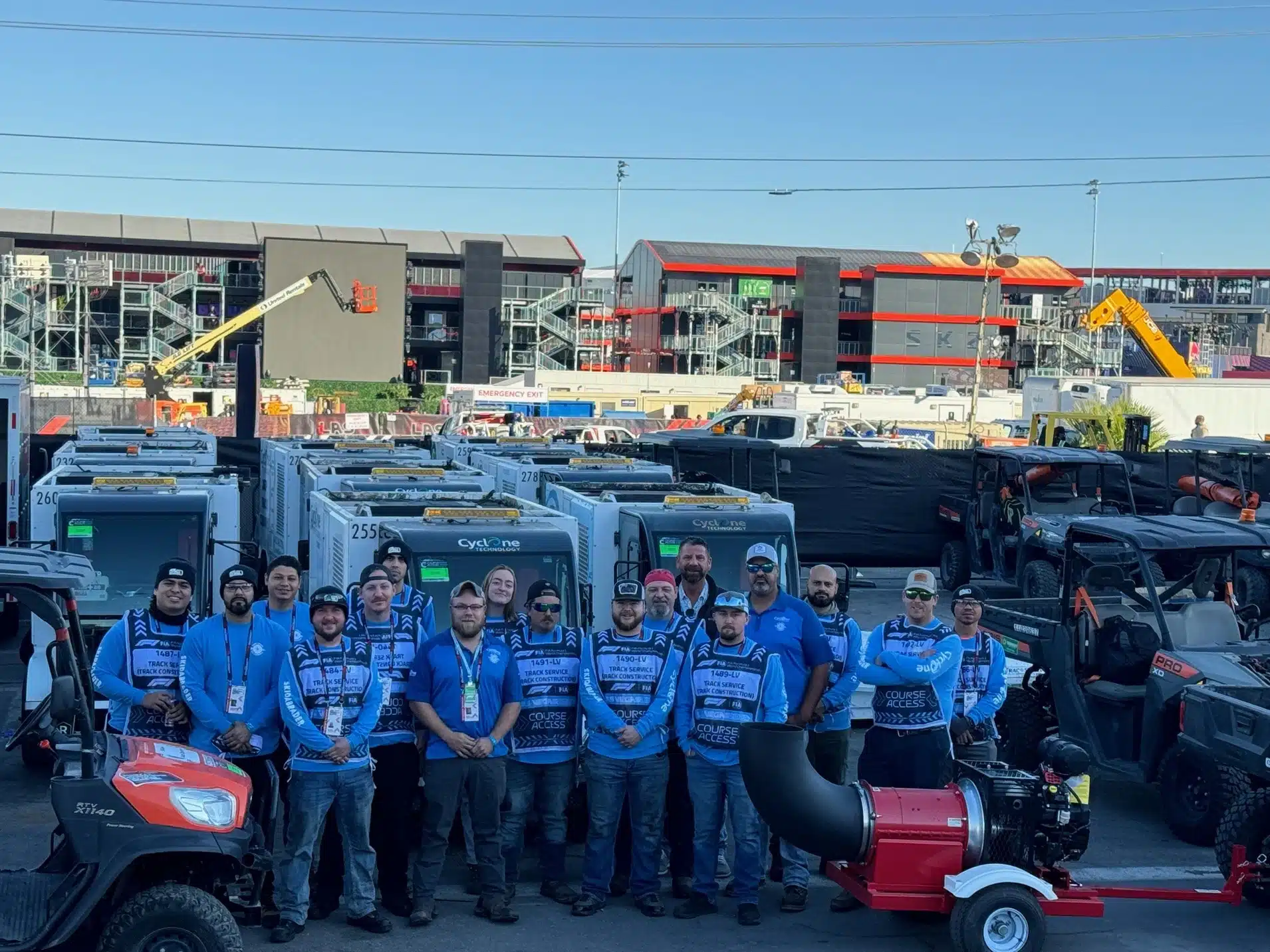 A group Skidabrader employees wearing matching blue shirts pose in front of various construction vehicles and equipment at an outdoor venue.