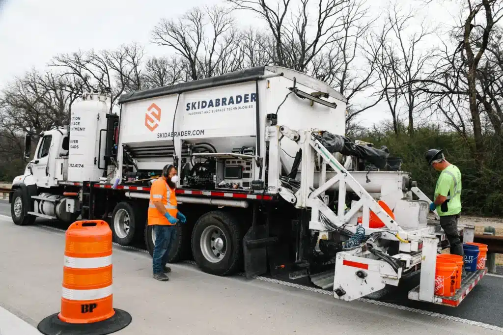 A truck with the branding "SKIDABRADER" is parked on a roadway. Two workers are operating equipment on the truck, wearing safety gear. Road construction barriers are visible in the foreground and trees are in the background.