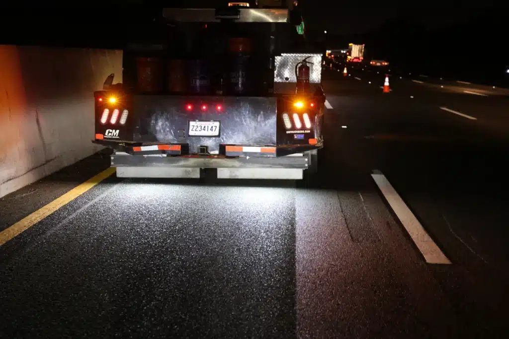A work truck with illuminated lights is parked on a dimly lit highway at night. Traffic cones are visible ahead, suggesting roadwork.