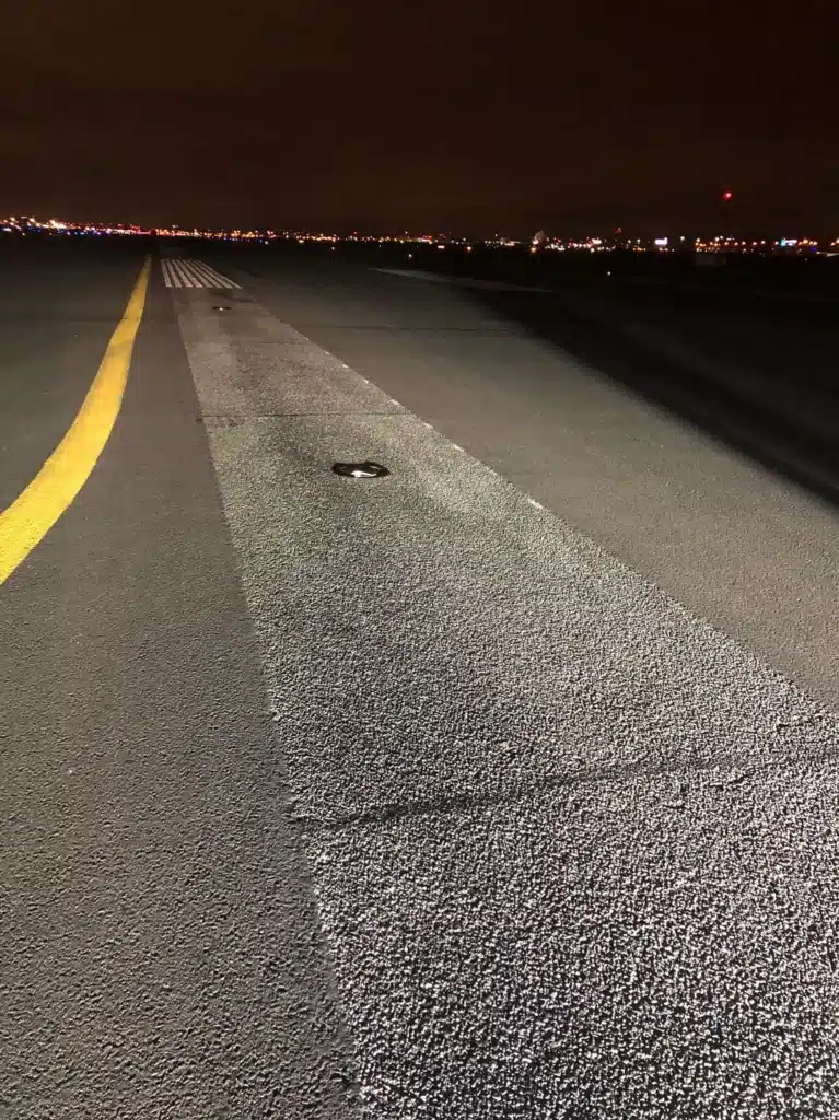 A runway at night with yellow markings and distant city lights in the background.
