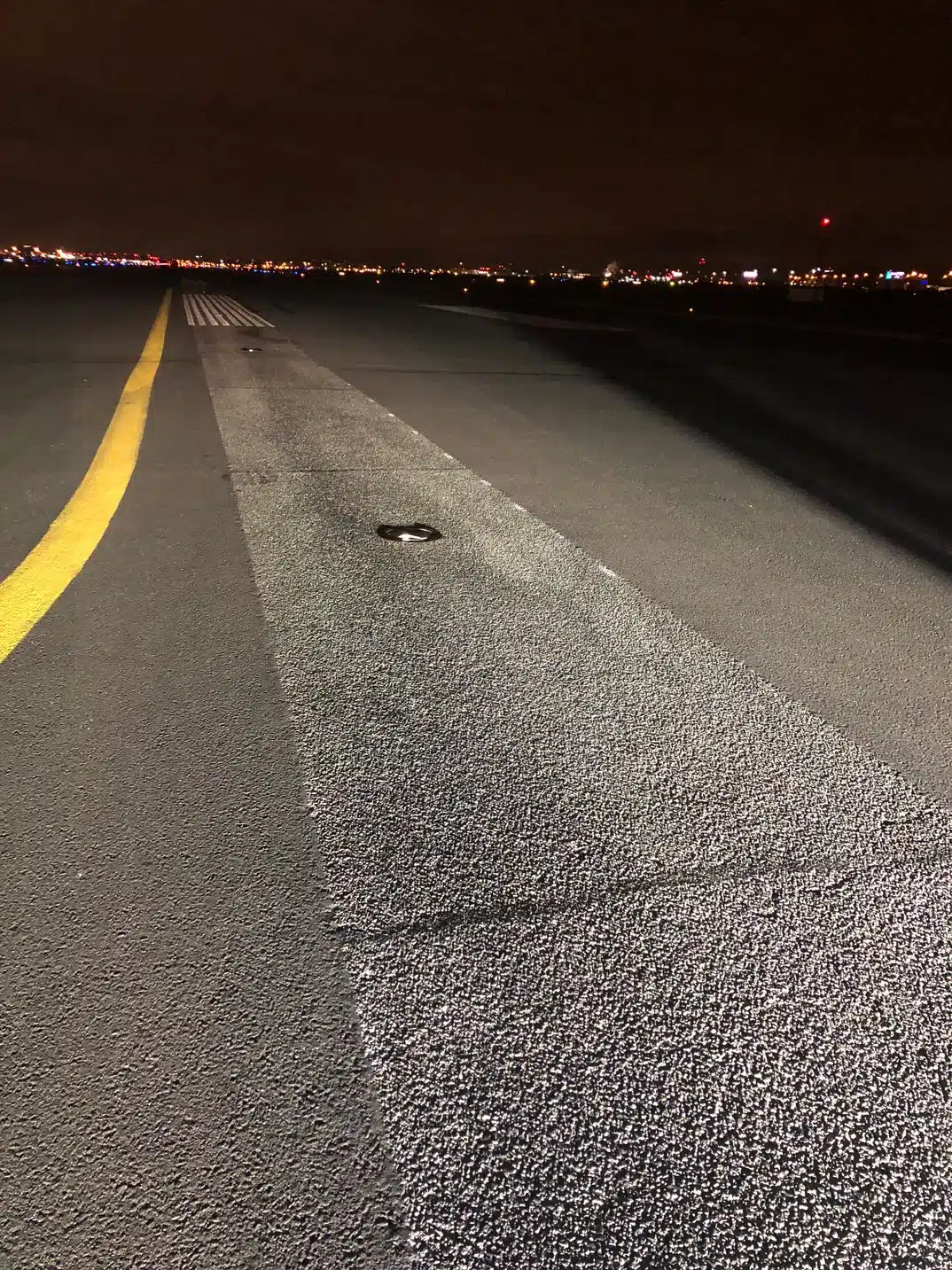 A runway at night with yellow markings and distant city lights in the background.