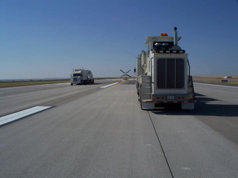 A row of trucks on a runway, with one truck facing forward and another truck in the background near a runway marking. Clear blue sky above.