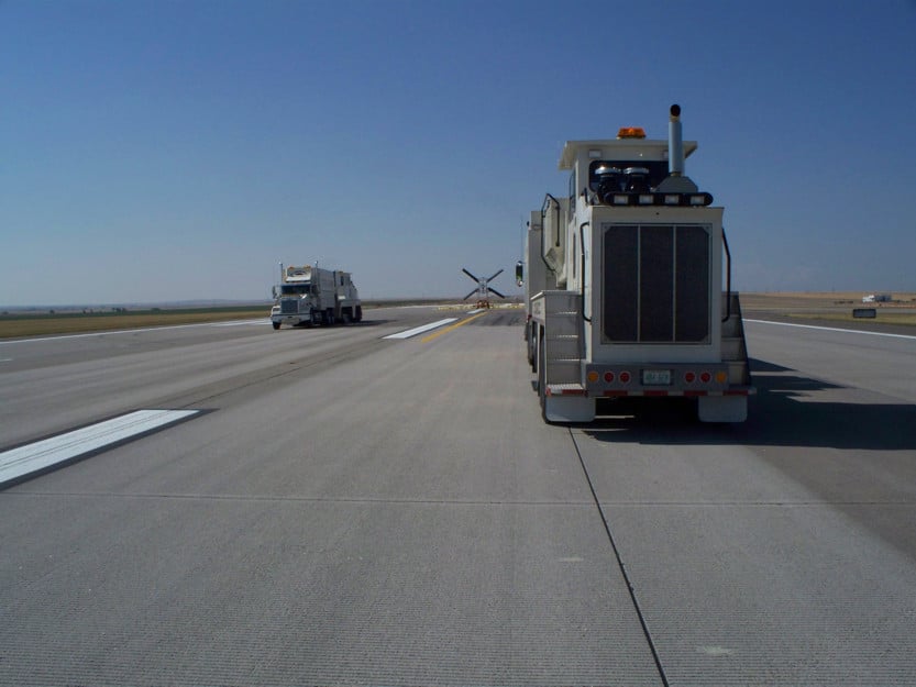 A row of trucks on a runway, with one truck facing forward and another truck in the background near a runway marking. Clear blue sky above.