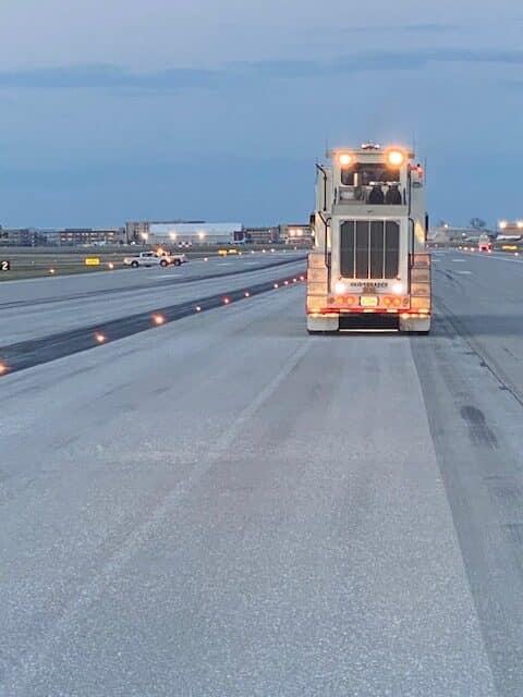 Runway view at dusk showing a large maintenance vehicle equipped with bright lights, moving away. The sky is darkening, creating a calm atmosphere.