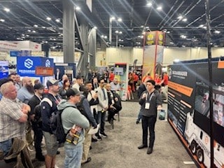 A presenter speaks to an attentive crowd at a trade show, with exhibition booths and bright displays in a large hall. The atmosphere is professional and focused.