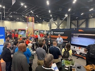 A group of people attentively watches a presentation at a trade show booth. The speaker stands near a large screen displaying information. The setting is busy and professional.