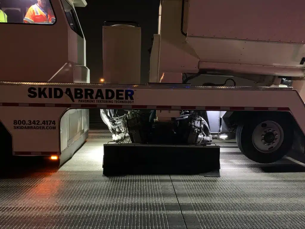 A Skidabrader vehicle parked at night, used for pavement texturing. It's illuminated from below, with a worker in a reflective jacket visible inside.